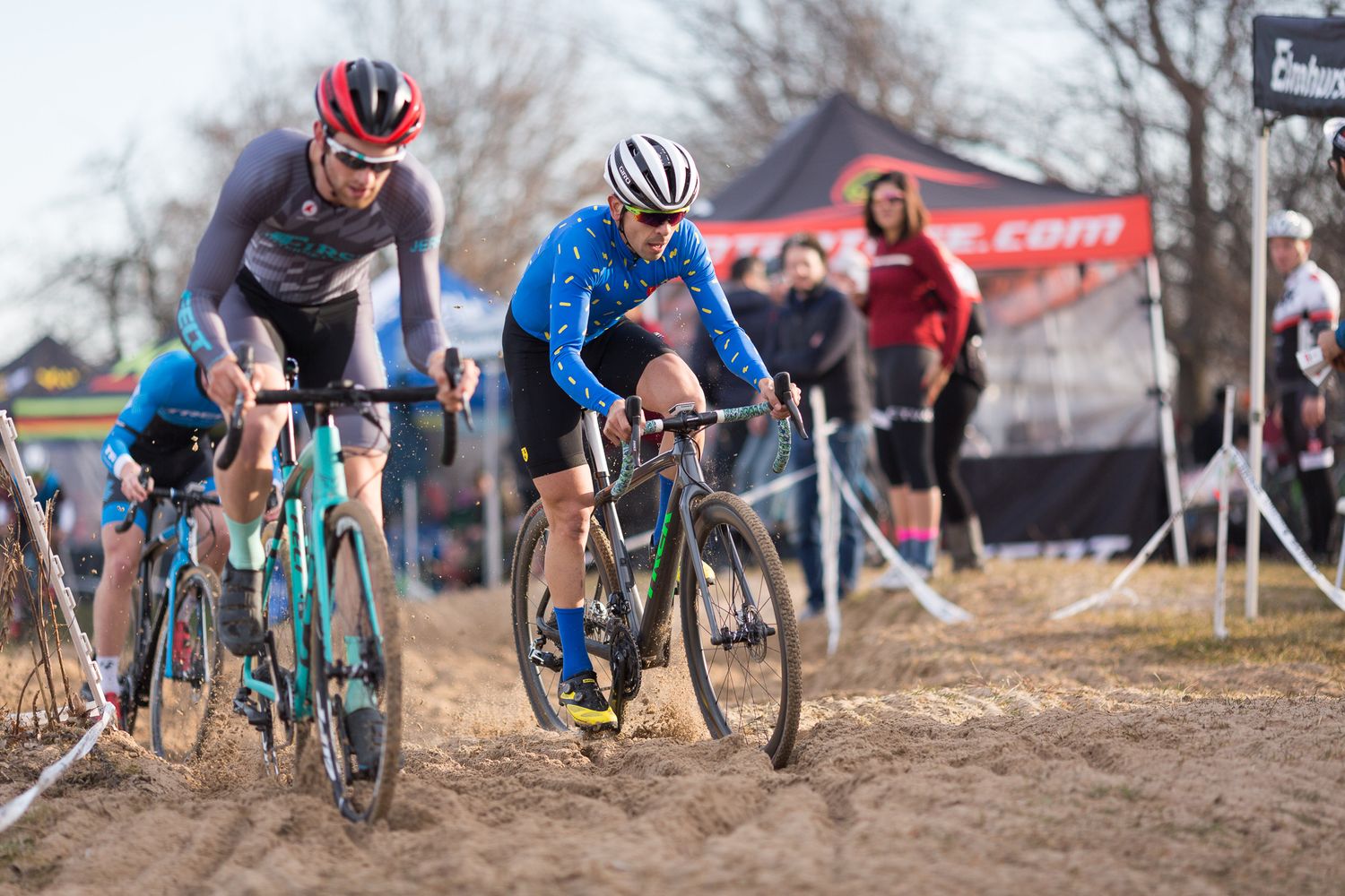 Two cyclocross riders navigate a sandy section during a competitive cycling race outdoors.