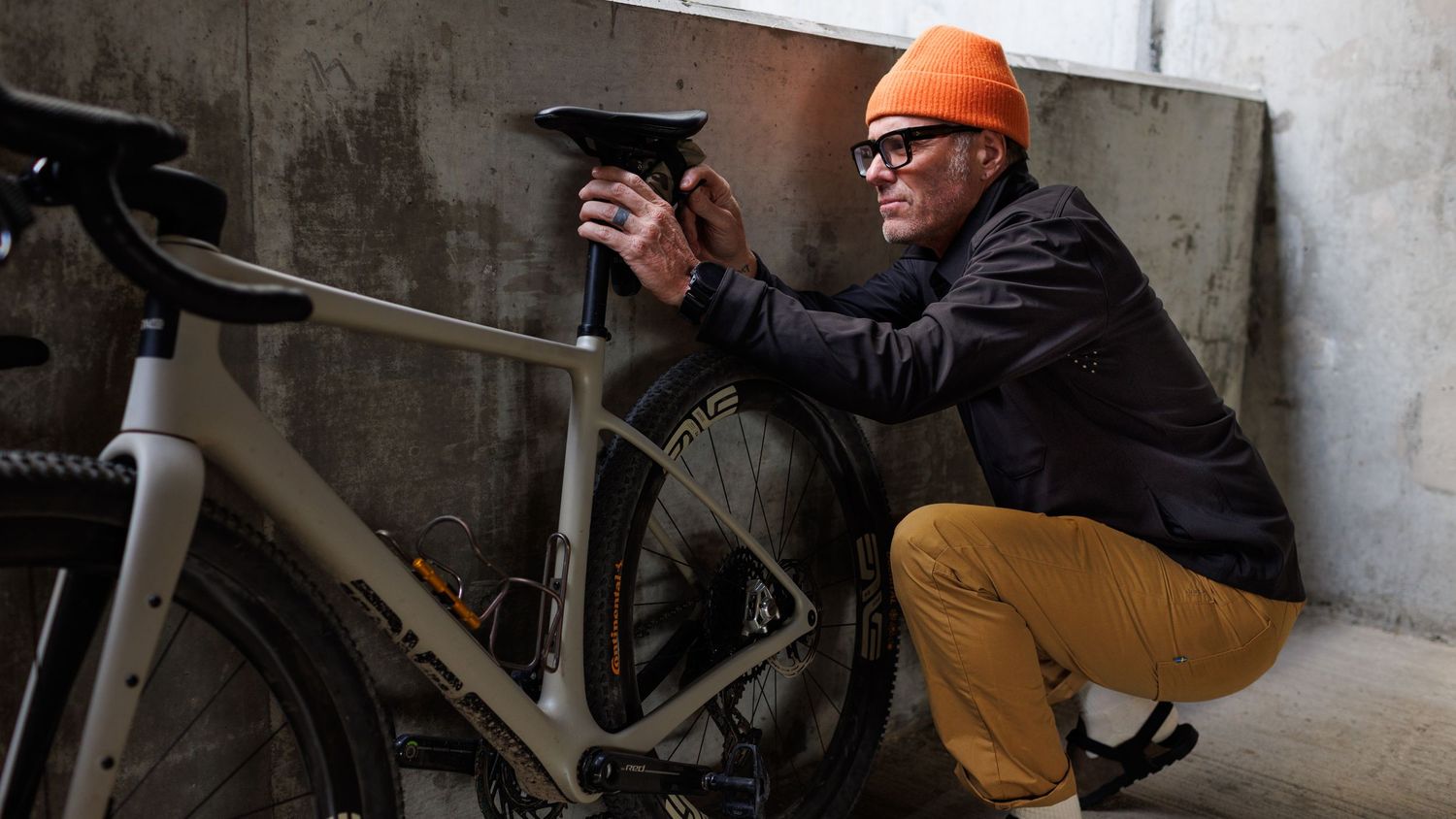 A man wearing an orange beanie and glasses crouching beside a gravel bike adjusting the saddle indoors.