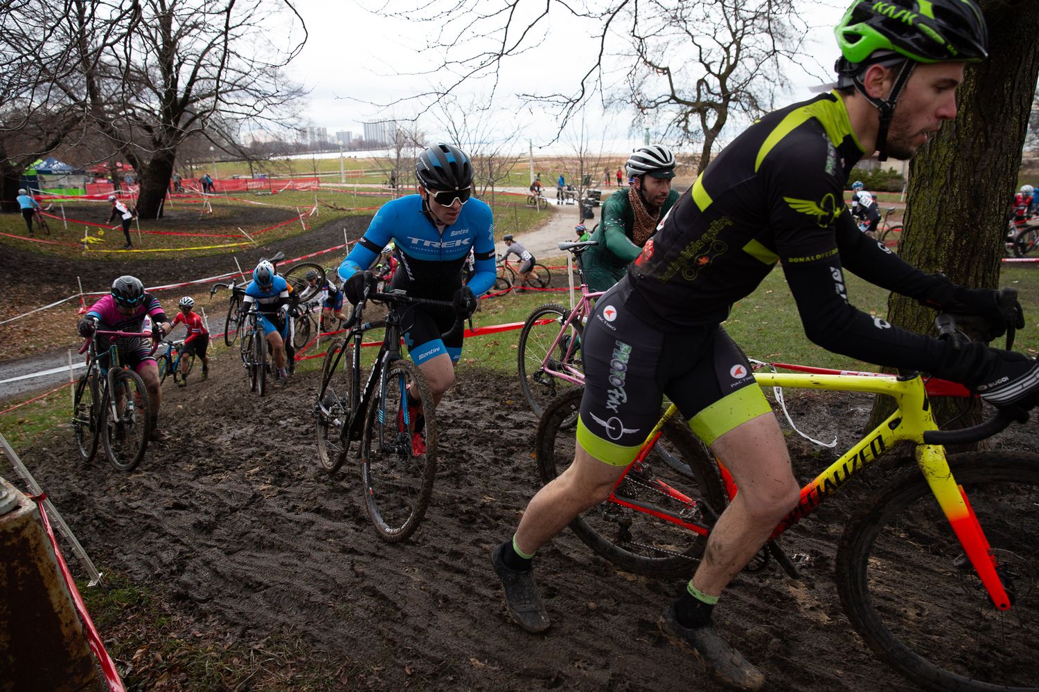 Muddy cyclocross race with cyclists navigating a wet, churned-up course through a park in cold weather.