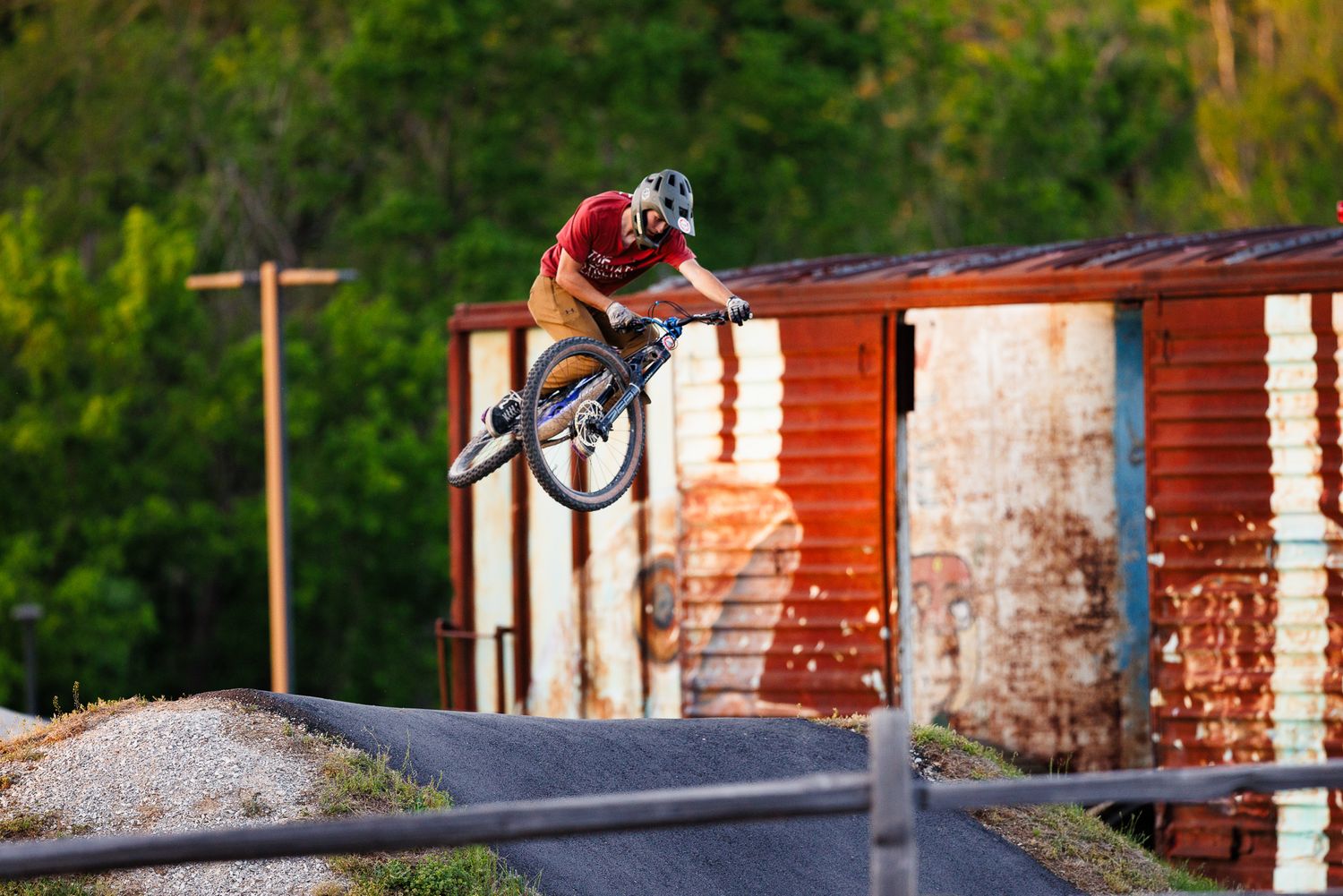 Mountain biker catches air off a jump near rustic metal buildings at a dirt bike park.