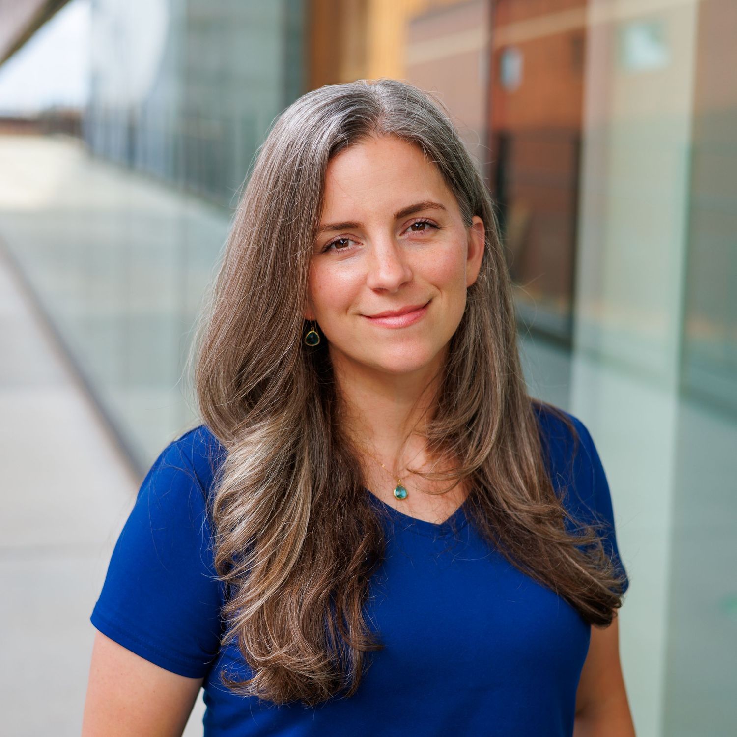 Professional headshot of a woman with long brown hair in a blue shirt outdoors.