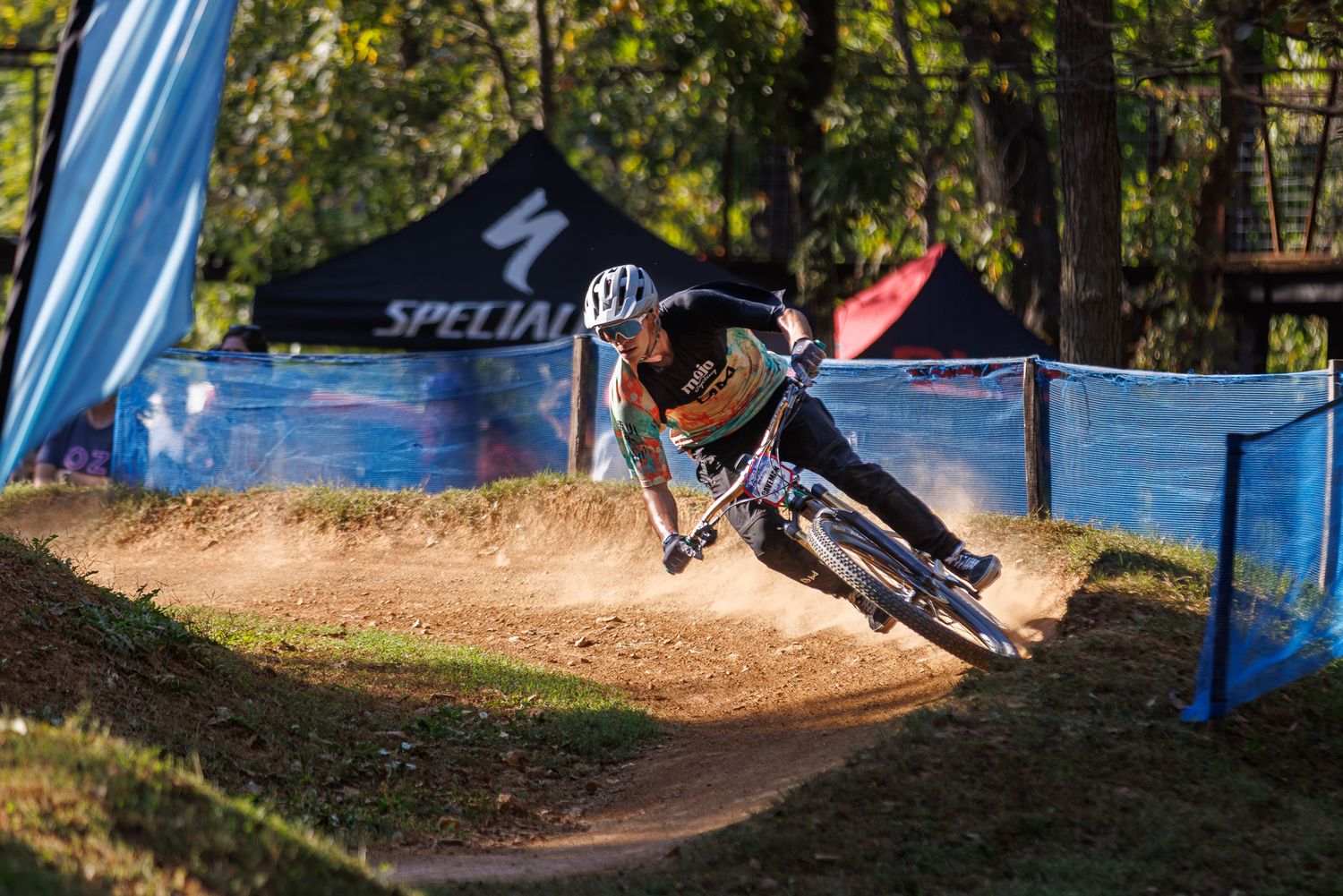 Mountain biker in black gear carving a sharp turn on a sandy dirt course at a Specialized event.