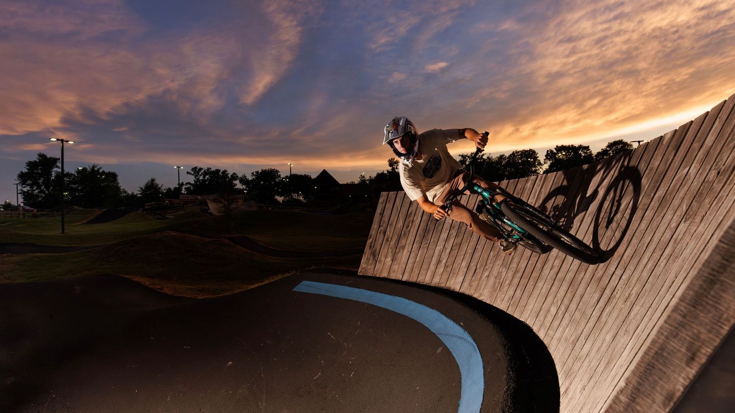BMX rider performing a high-speed turn on a wooden skate park ramp at sunset with dramatic cloudy sky.