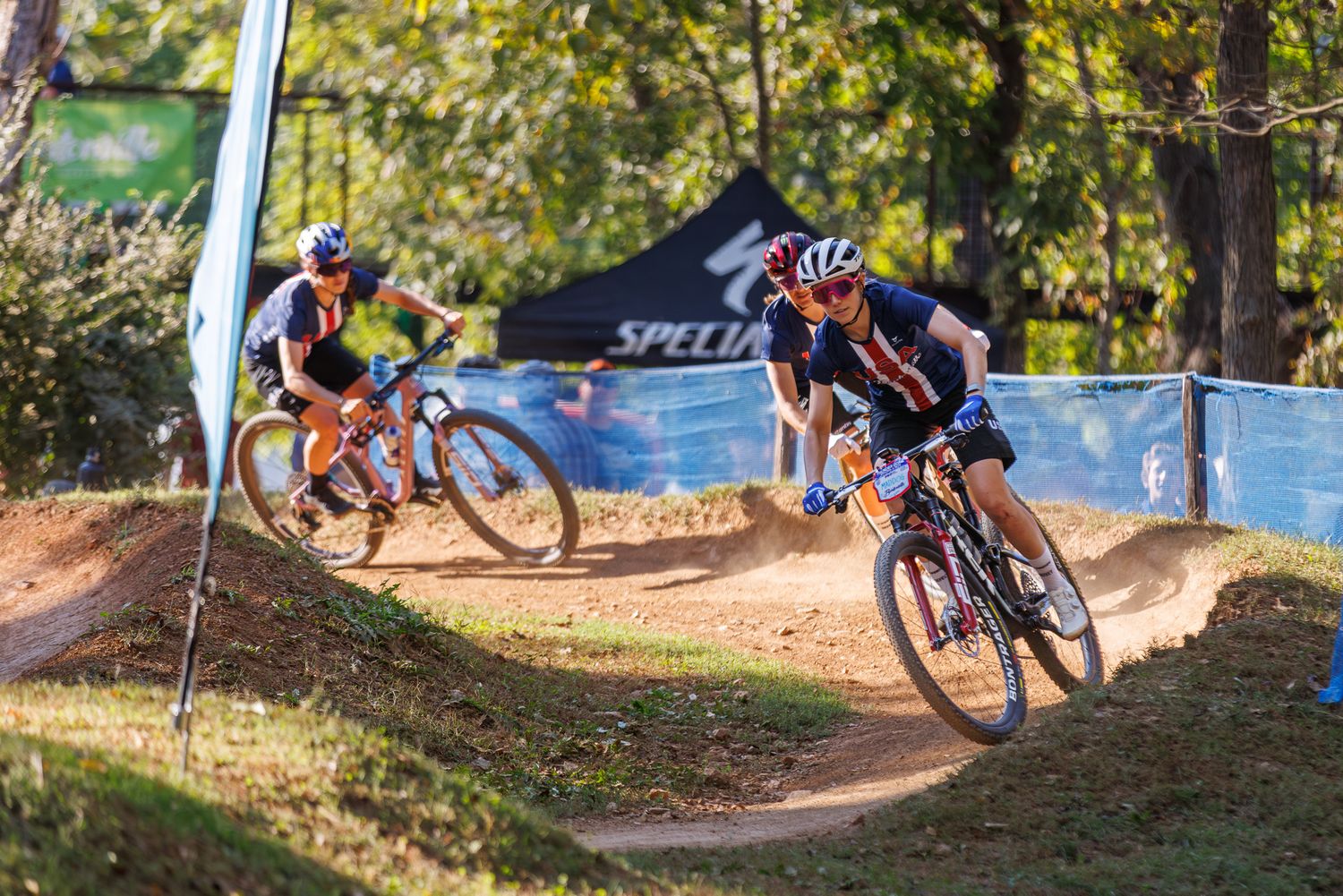 Two mountain bikers competing on a winding dirt trail near a Specialized sponsor banner.