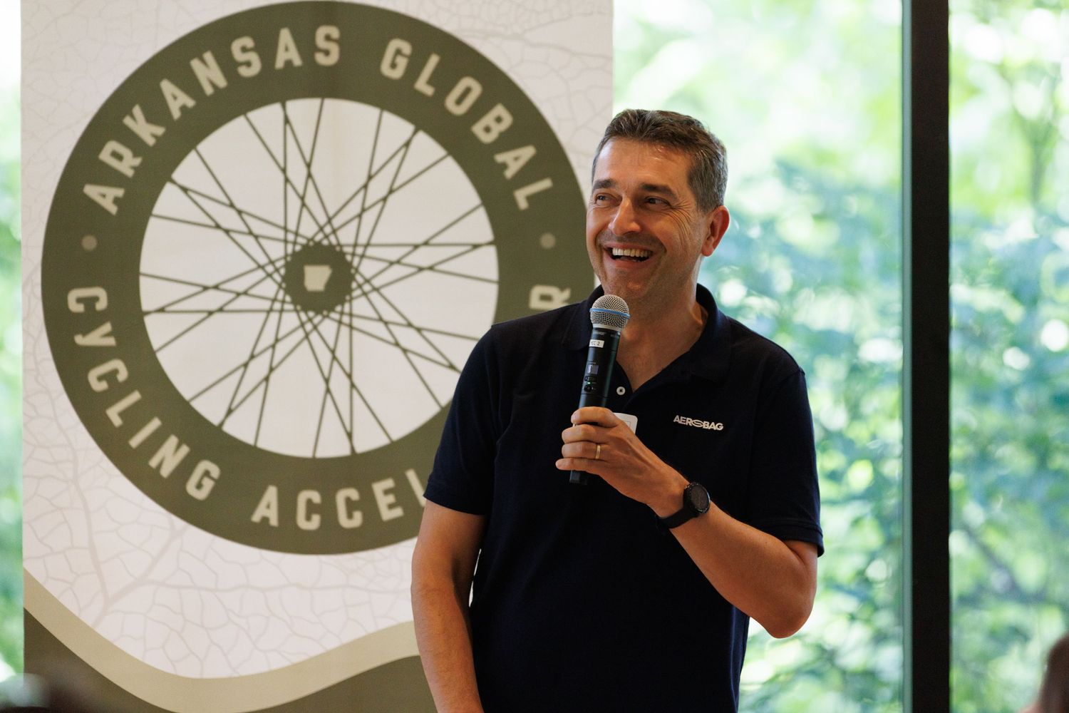 Man speaking into a microphone in front of an Arkansas Global Cycling Access banner.