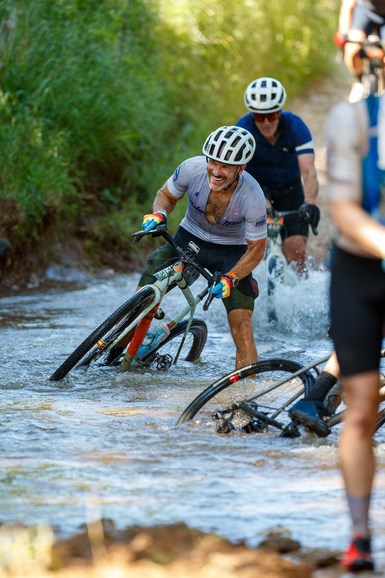 Cyclists pushing bikes through a rocky river crossing during a gravel bike race.