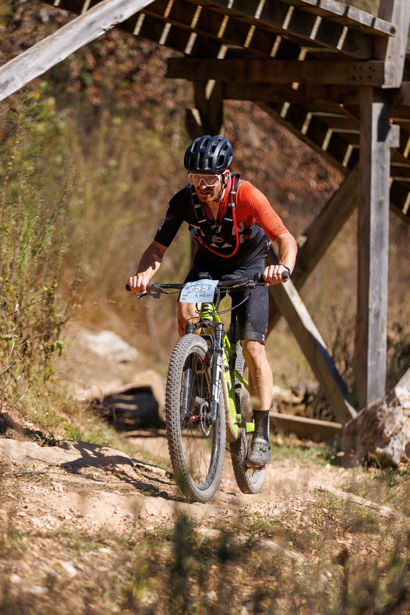 Male mountain biker in red and black jersey rides rocky trail beneath wooden bridge.