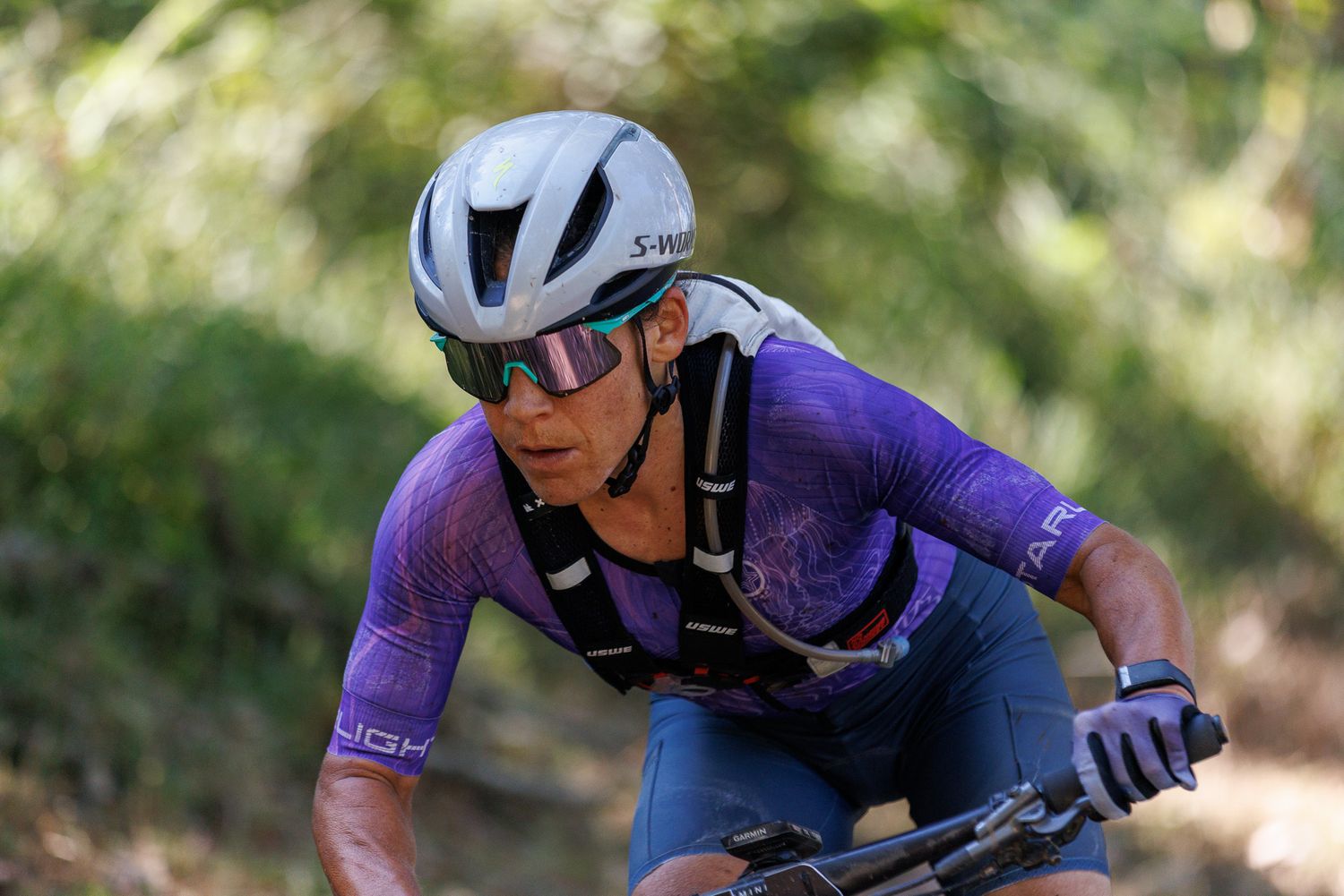 Close-up of focused female mountain biker in purple jersey wearing sunglasses on trail.