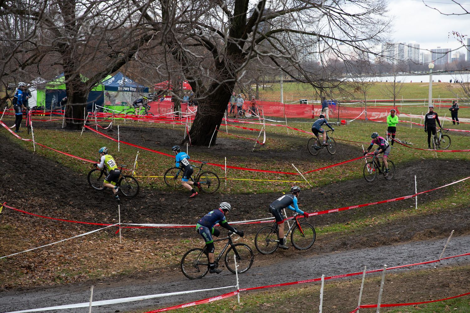 Aerial view of cyclocross racers navigating a muddy park course marked with red and white tape.