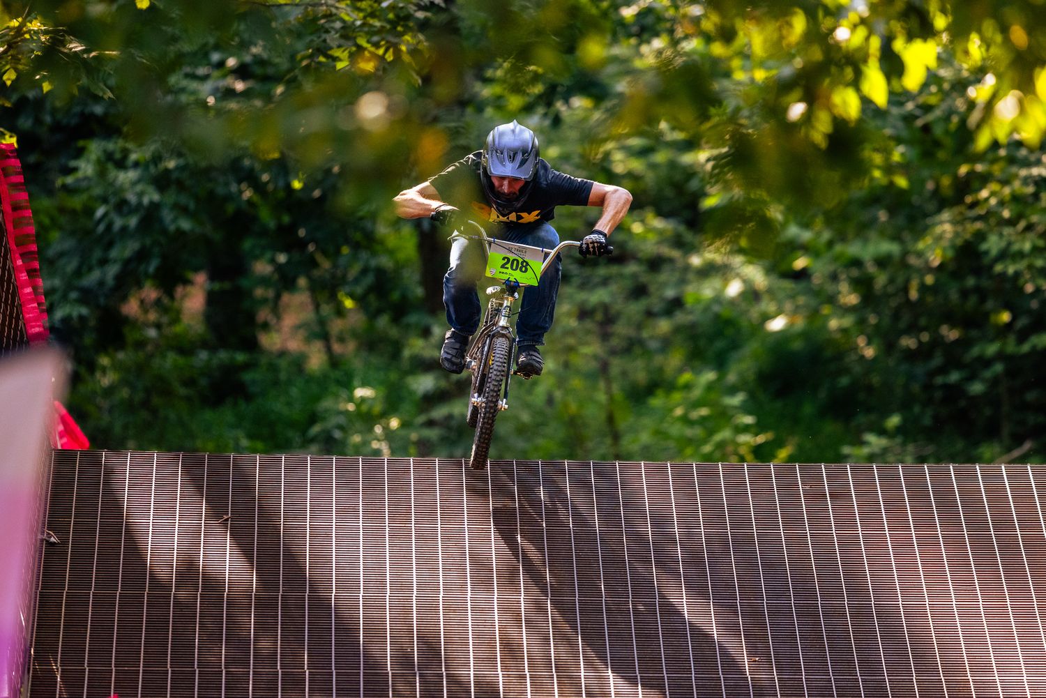 Young mountain biker launches off wooden jump ramp surrounded by lush green forest trees.
