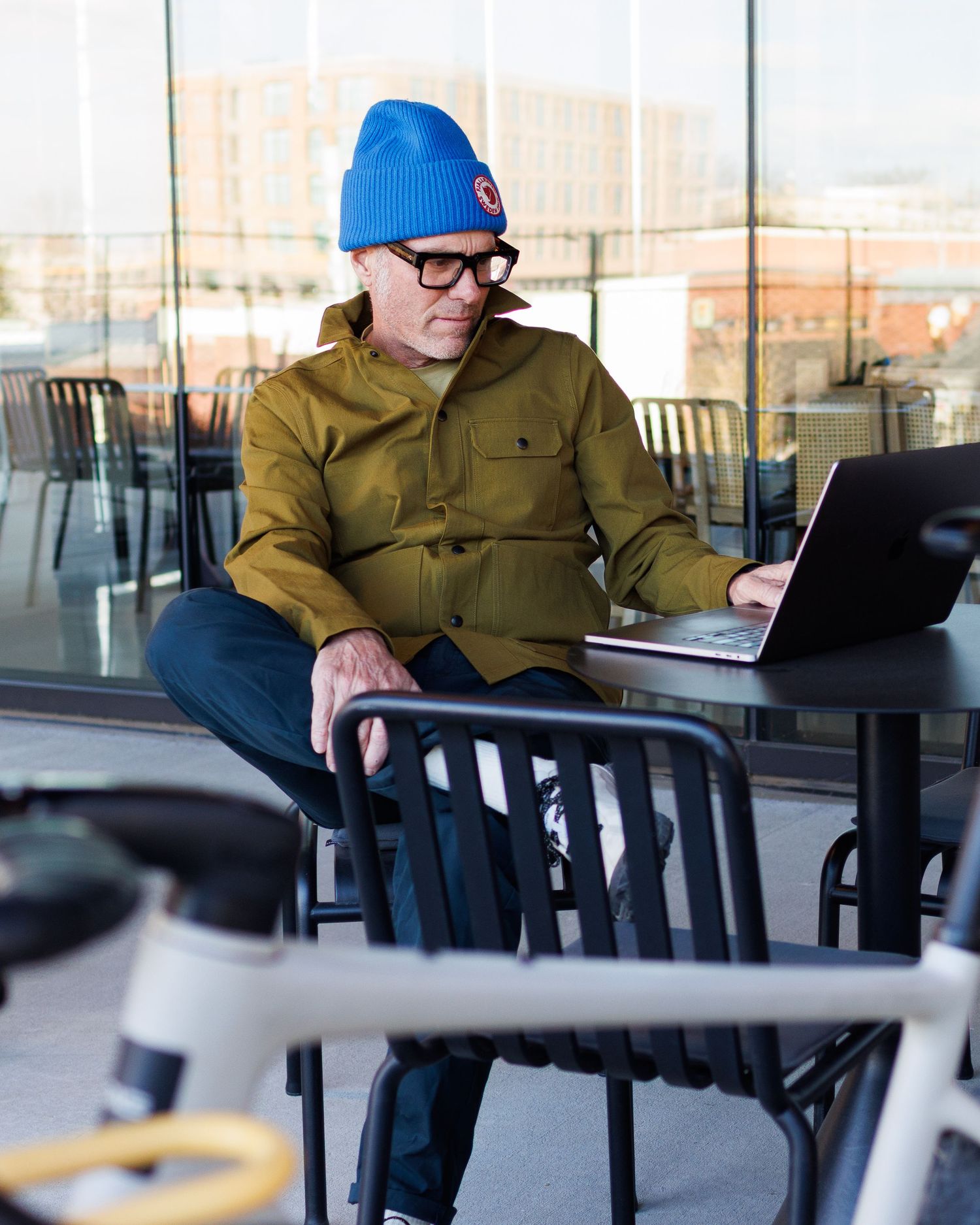Man in olive jacket and blue beanie working on laptop at outdoor cafe with bike nearby.