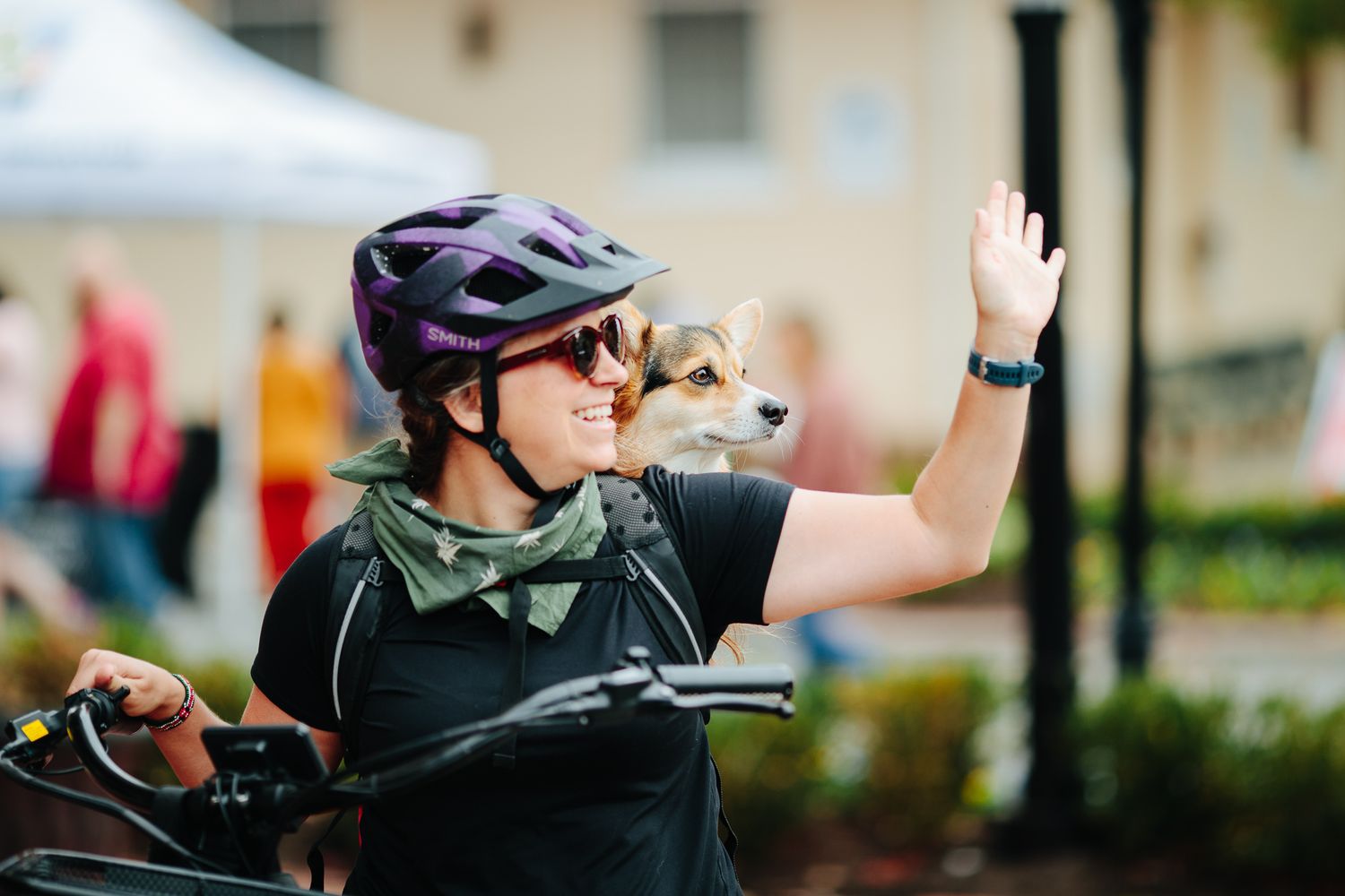 A smiling woman wearing a purple bike helmet carries a small dog while waving outdoors.