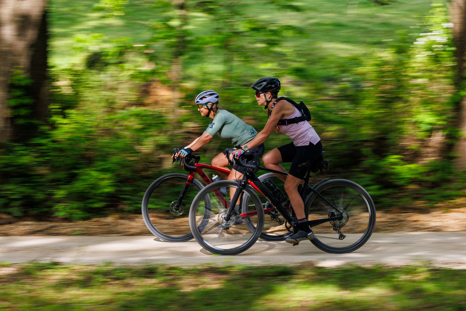 Two cyclists ride road bikes side by side on a tree-lined path with motion blur background.
