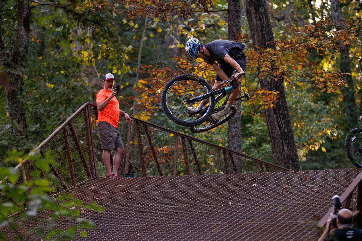 Cyclist performs big air trick off wooden ramp feature as spectator watches.