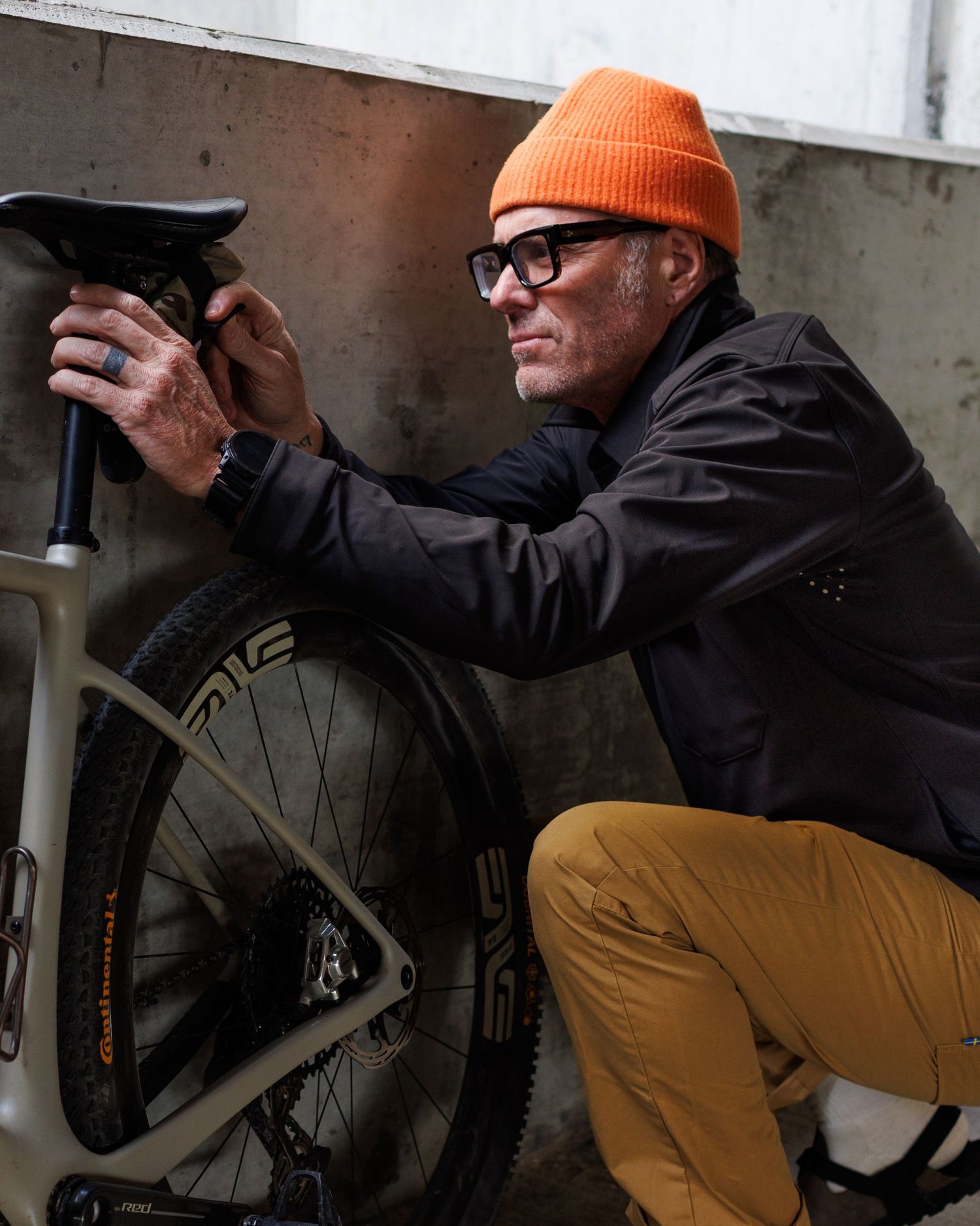 Man in orange beanie and black jacket examining bicycle outdoors.