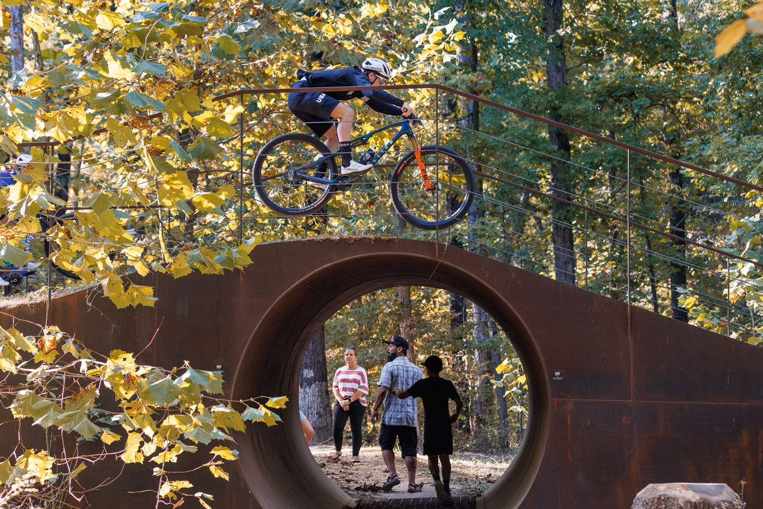 Mountain biker performing a jump trick viewed through a large circular metal frame in a forest.