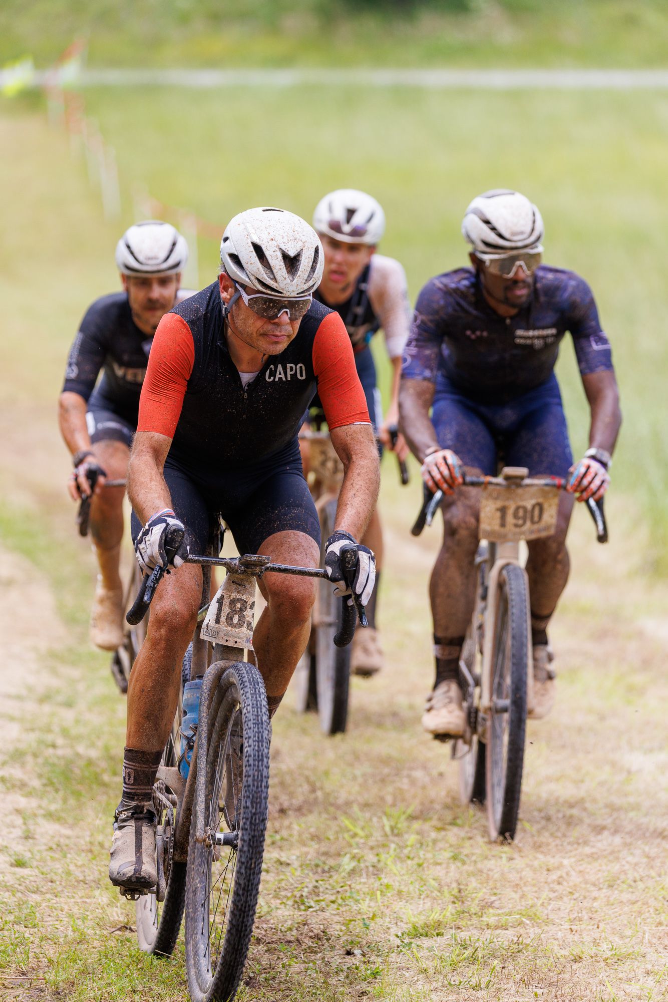 Group of muddy gravel cyclists led by a rider in a Capo jersey competing in an off-road race.