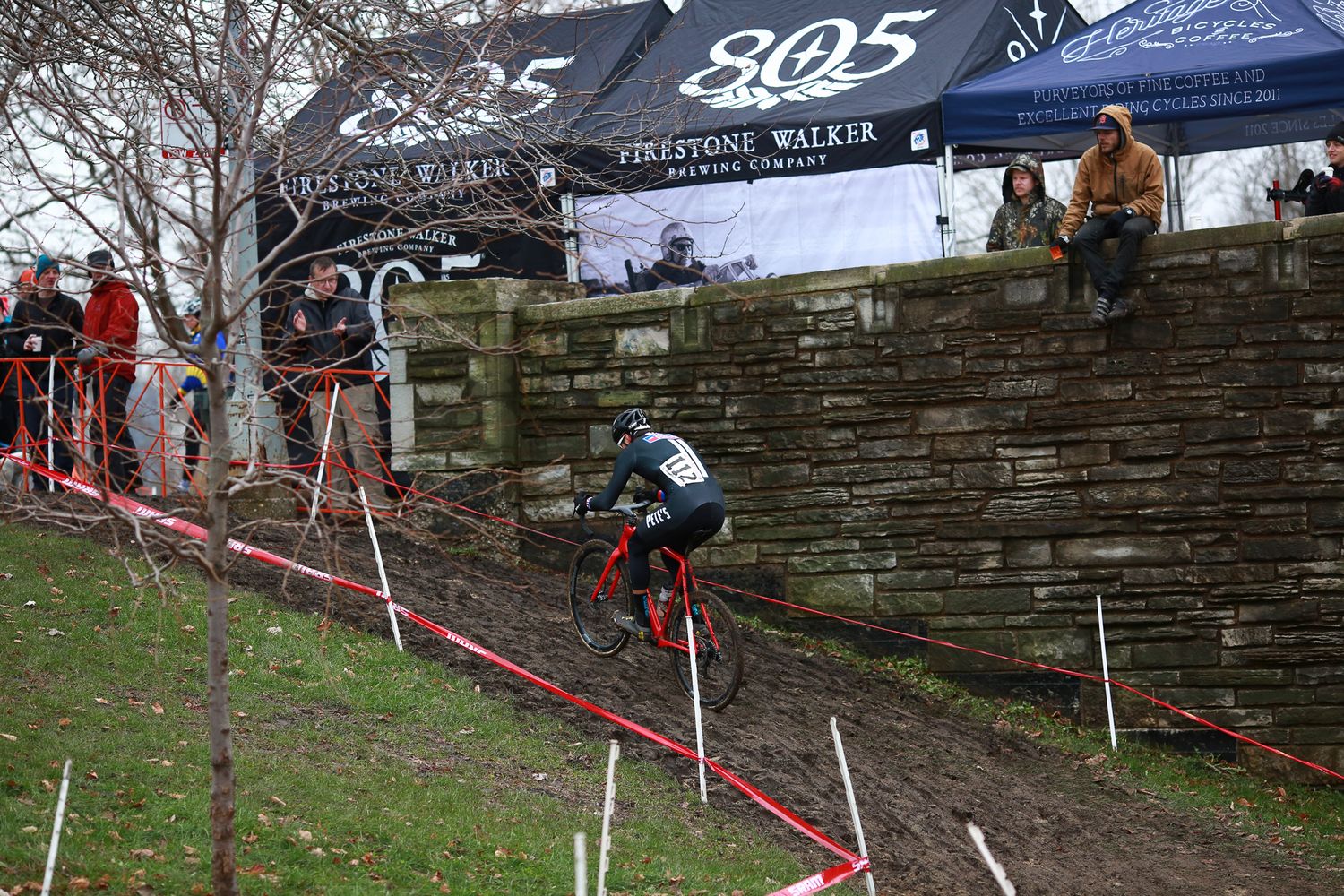 Cyclocross racer descends a steep hill beside a stone wall with spectators watching from above.