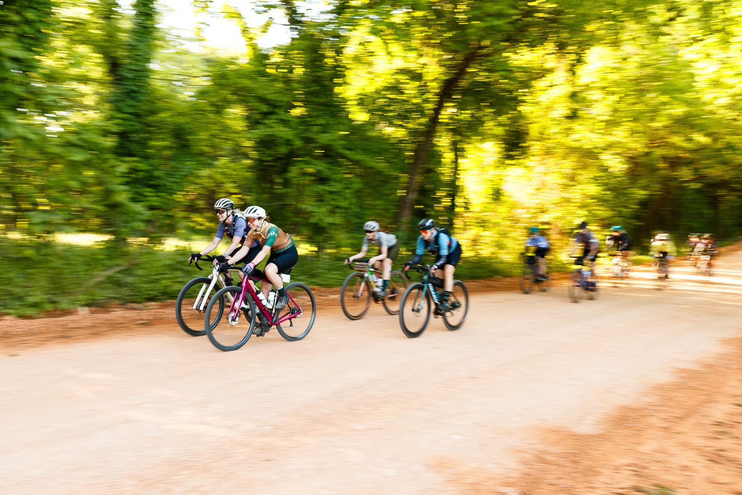 Motion-blurred group of cyclists racing on a dirt road through a lush green forest.