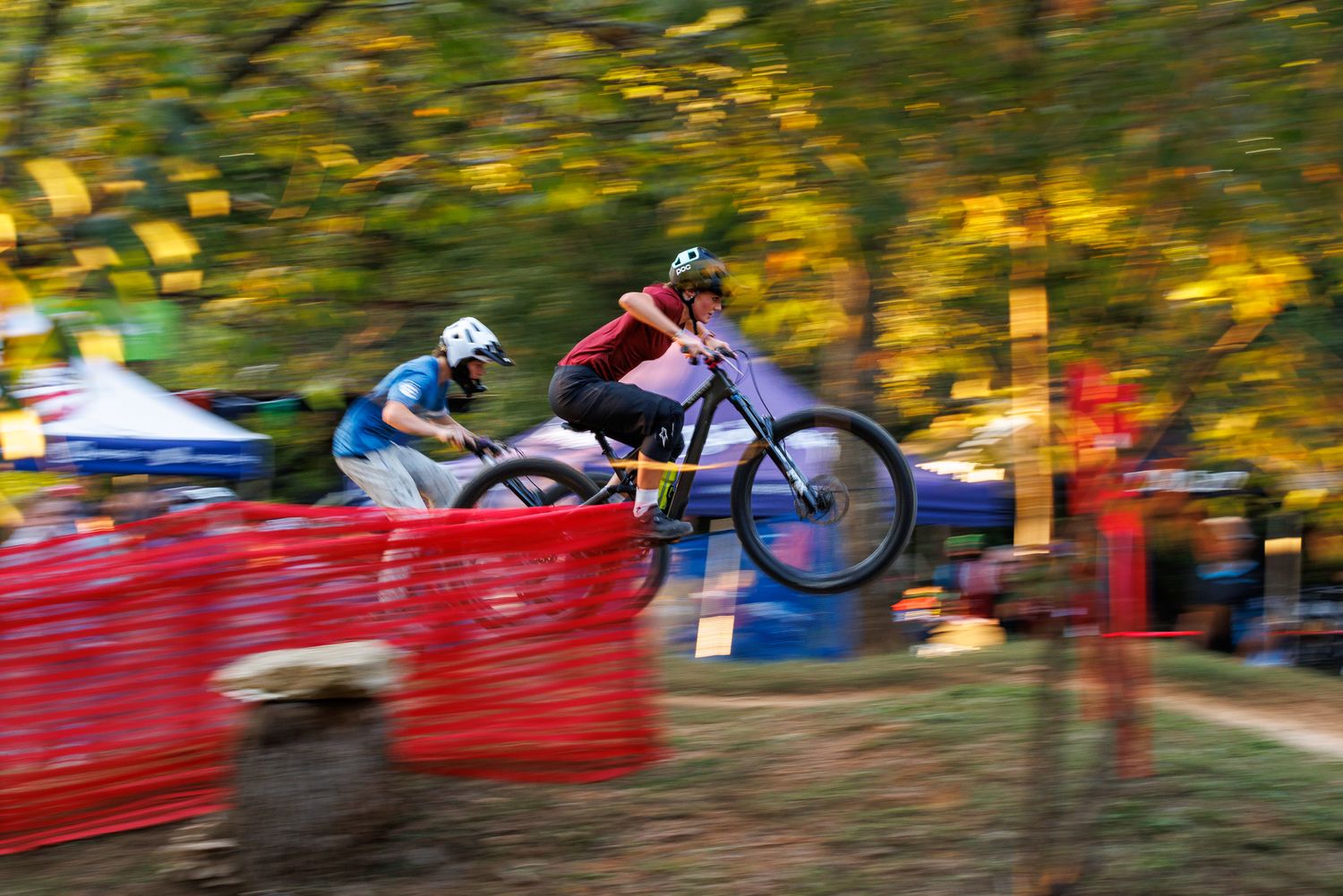 Motion-blurred mountain biker launches over red barrier at autumn MTB race.