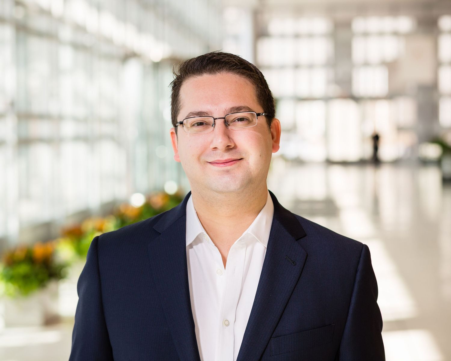 Smiling man in glasses and navy blazer posing in a bright modern lobby.