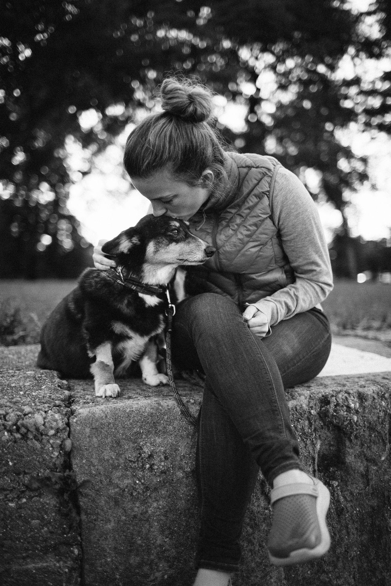 Woman kissing her Corgi dog while sitting on stone wall outdoors, black and white photo.
