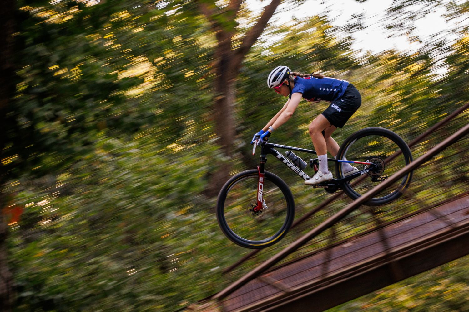 Mountain biker in blue jersey catches air on wooden ramp feature during race.