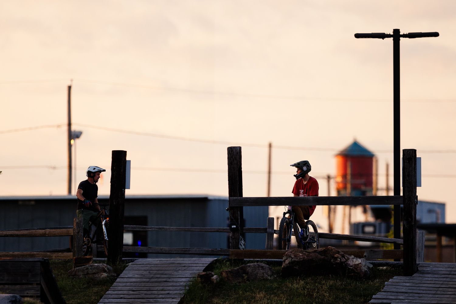 Two silhouetted mountain bikers stand on wooden trail bridges at dusk near a water tower.