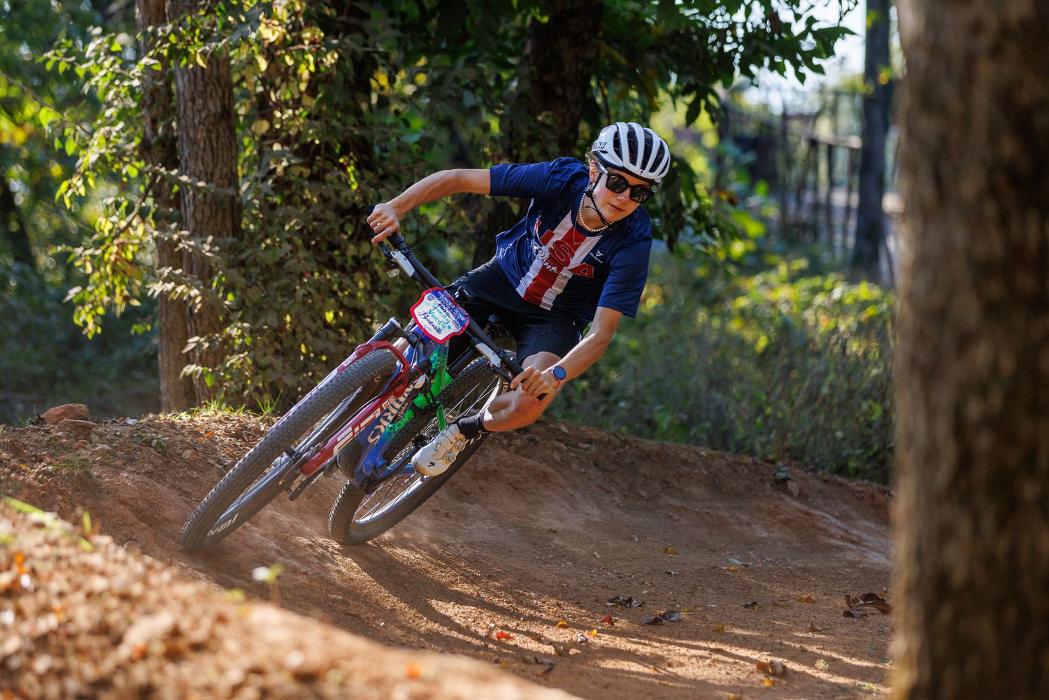 Mountain biker leaning into a banked dirt corner on a forest trail during a race.