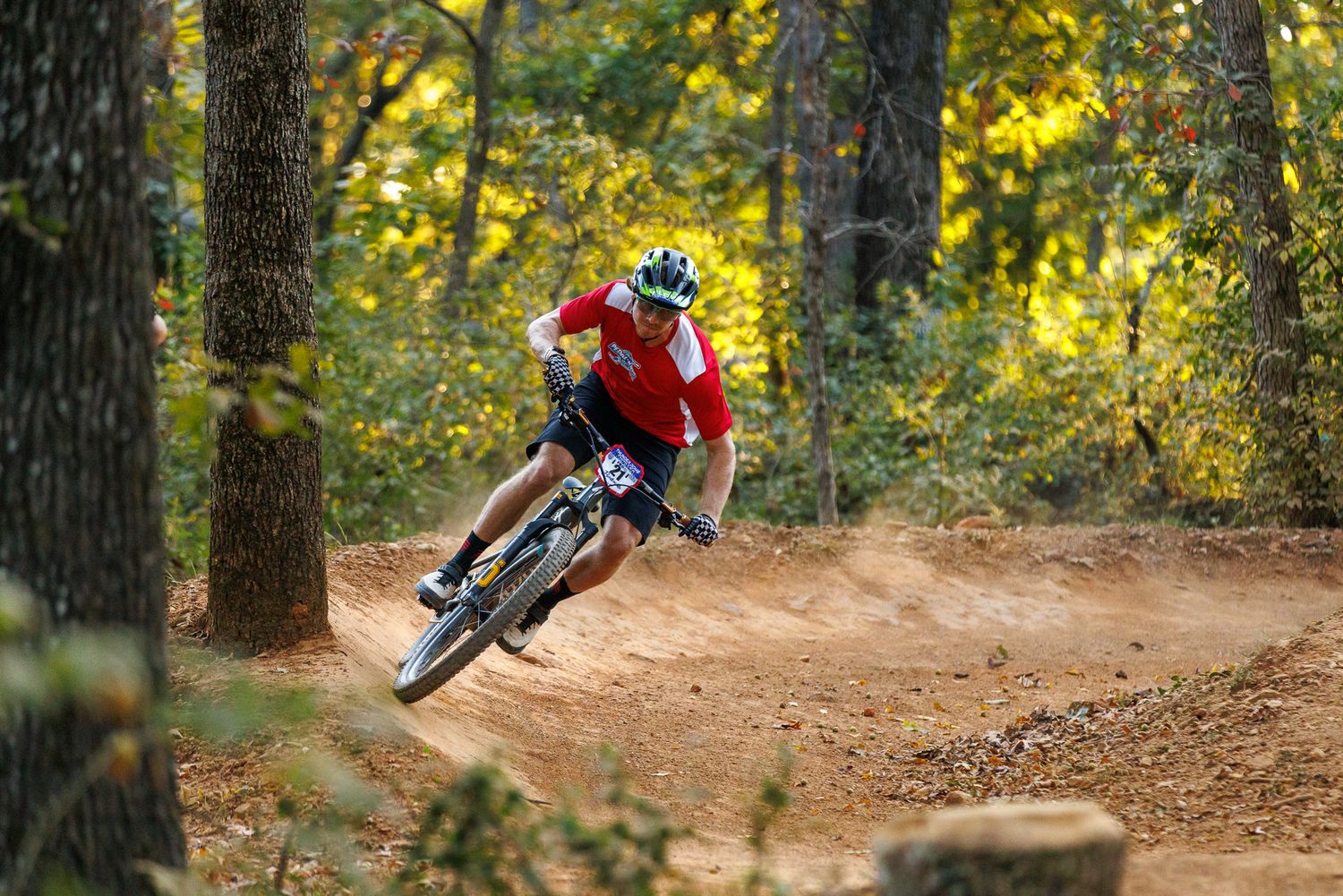 Mountain biker in red jersey leans into fast dirt corner among autumn forest trees.