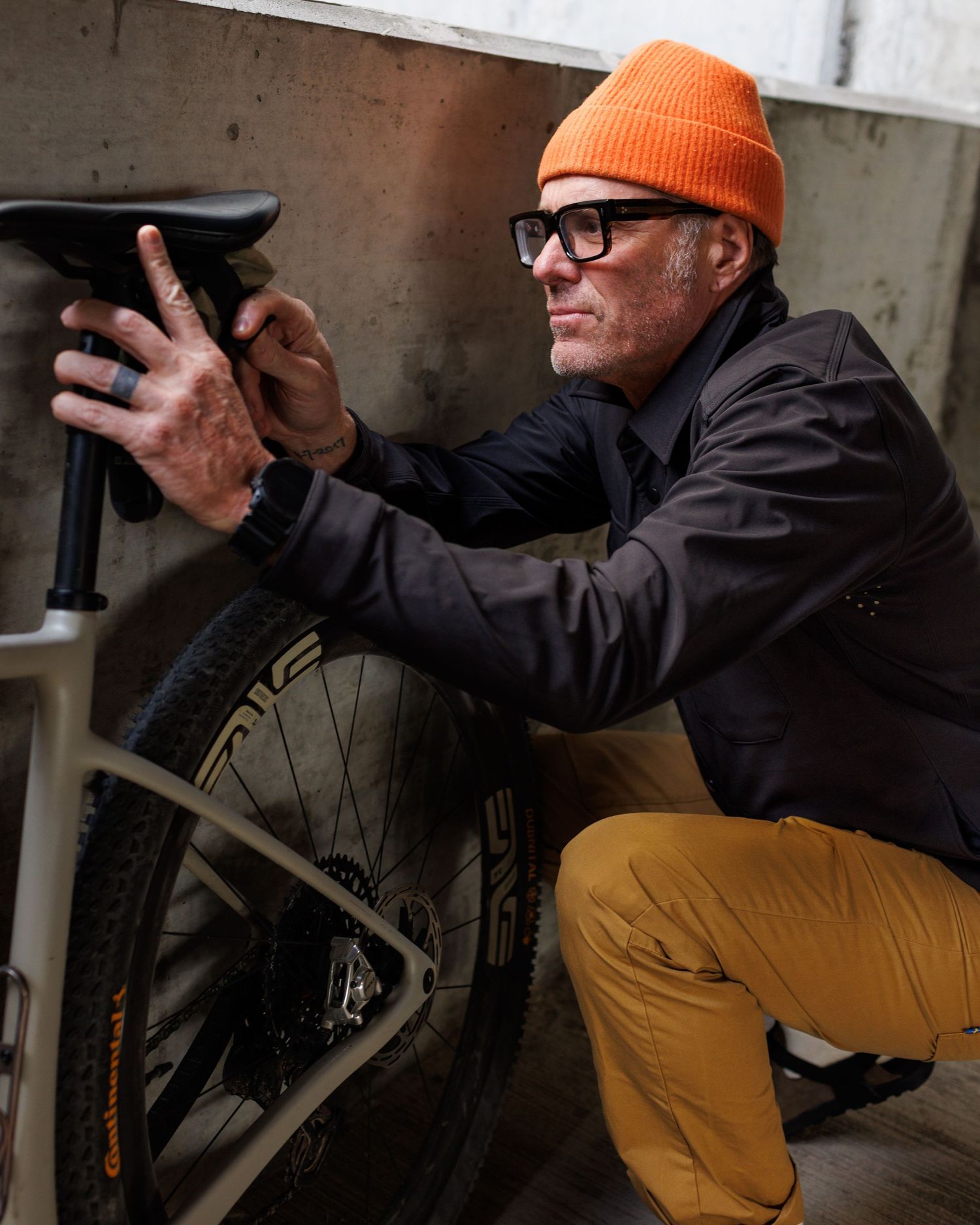 Man in orange beanie and glasses crouches to inspect bicycle wheel against concrete wall.