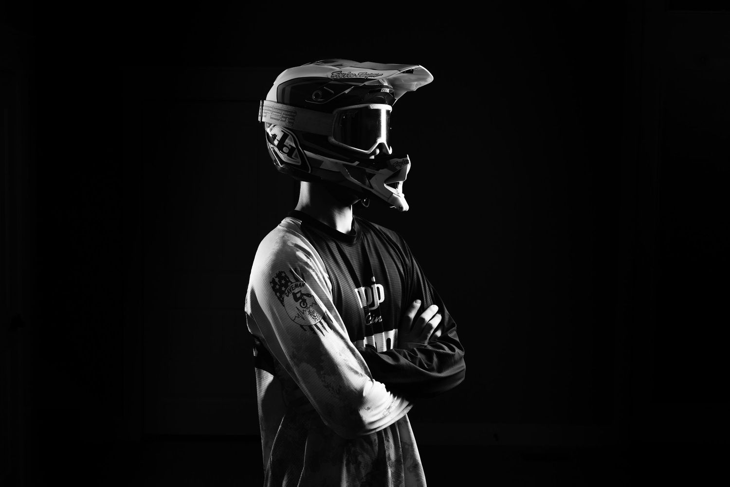 Black and white photo of a motocross rider in helmet and goggles standing with arms crossed against dark background.