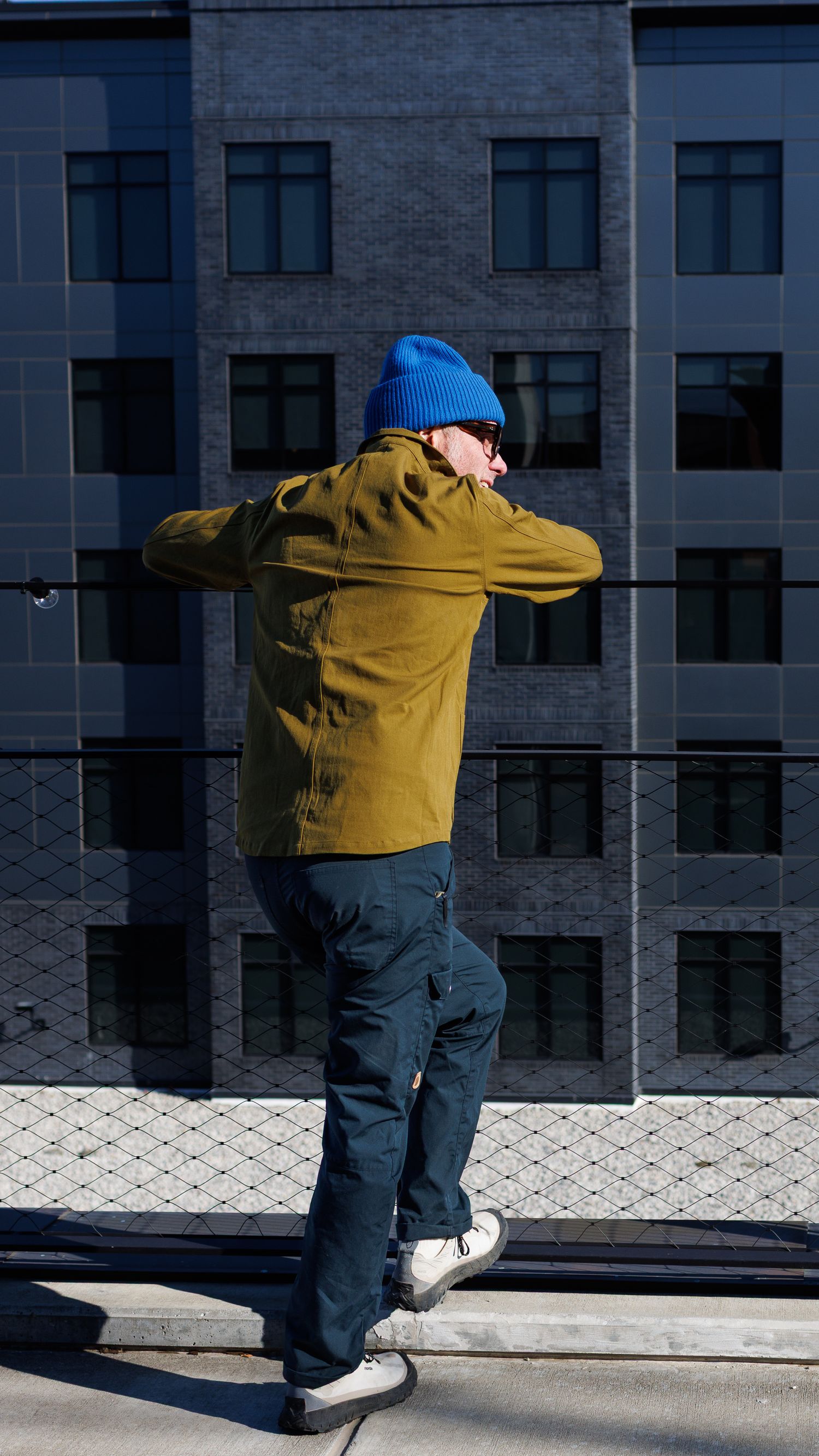 Person in olive jacket and blue beanie leaning on a railing with a modern building backdrop.