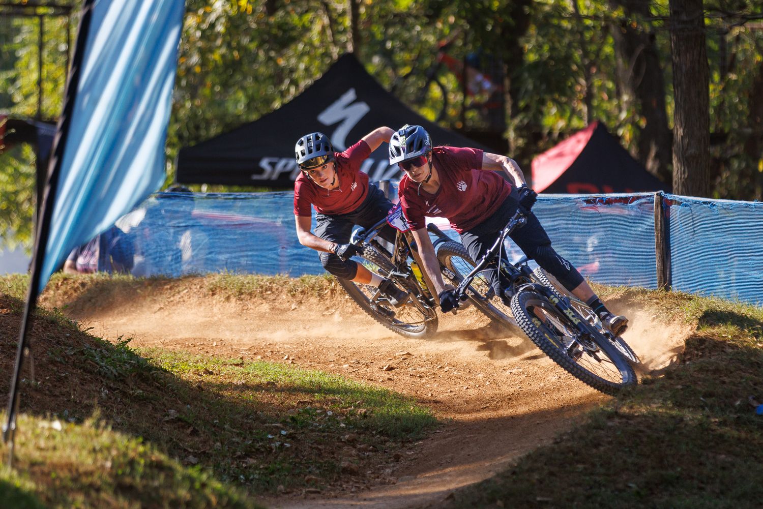 Two mountain bikers racing side by side on a dusty dirt track with Specialized banners in background.