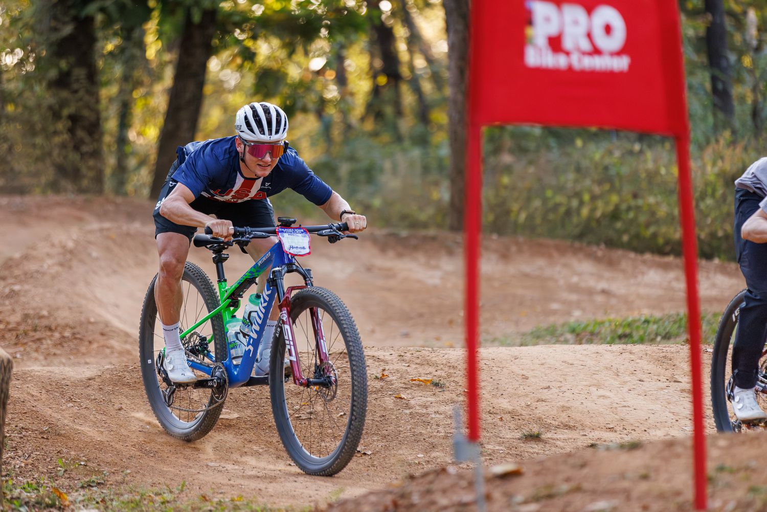 Male cyclist rides blue mountain bike past red Pro Bike Center banner on dirt trail.