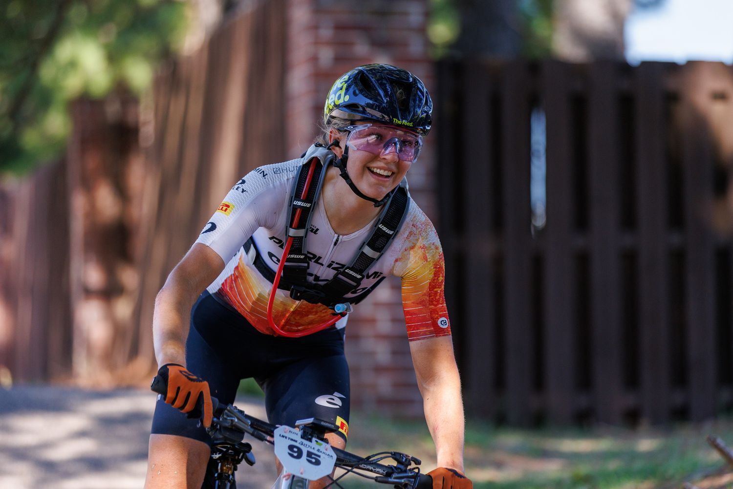 Smiling female cyclist in orange gradient jersey rides mountain bike past brick wall.