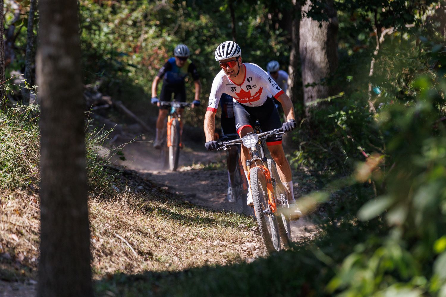Mountain biker in orange and white jersey leads group through dusty forest singletrack trail.