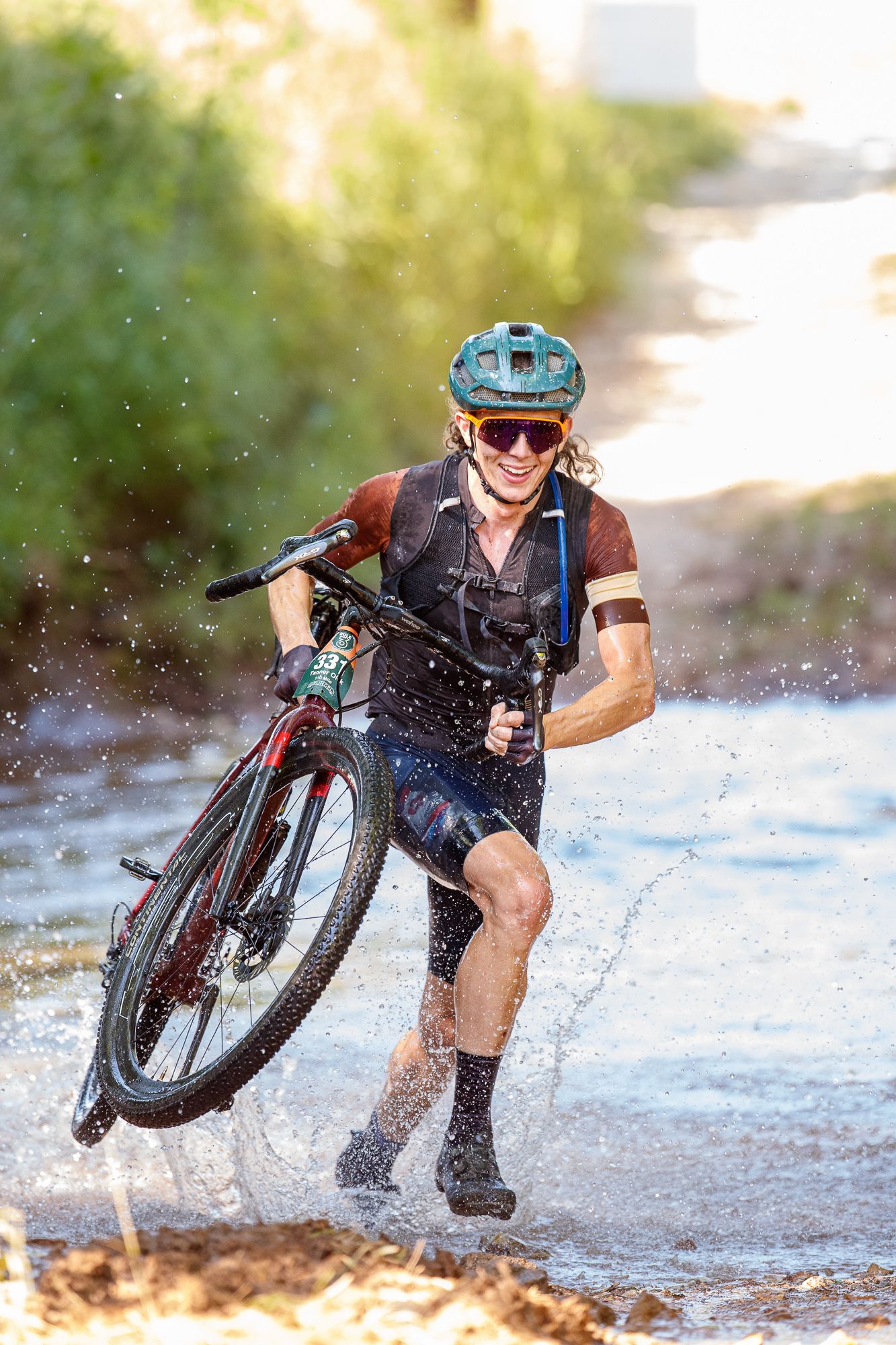 Smiling cyclist carrying mountain bike through splashing water stream on outdoor trail.