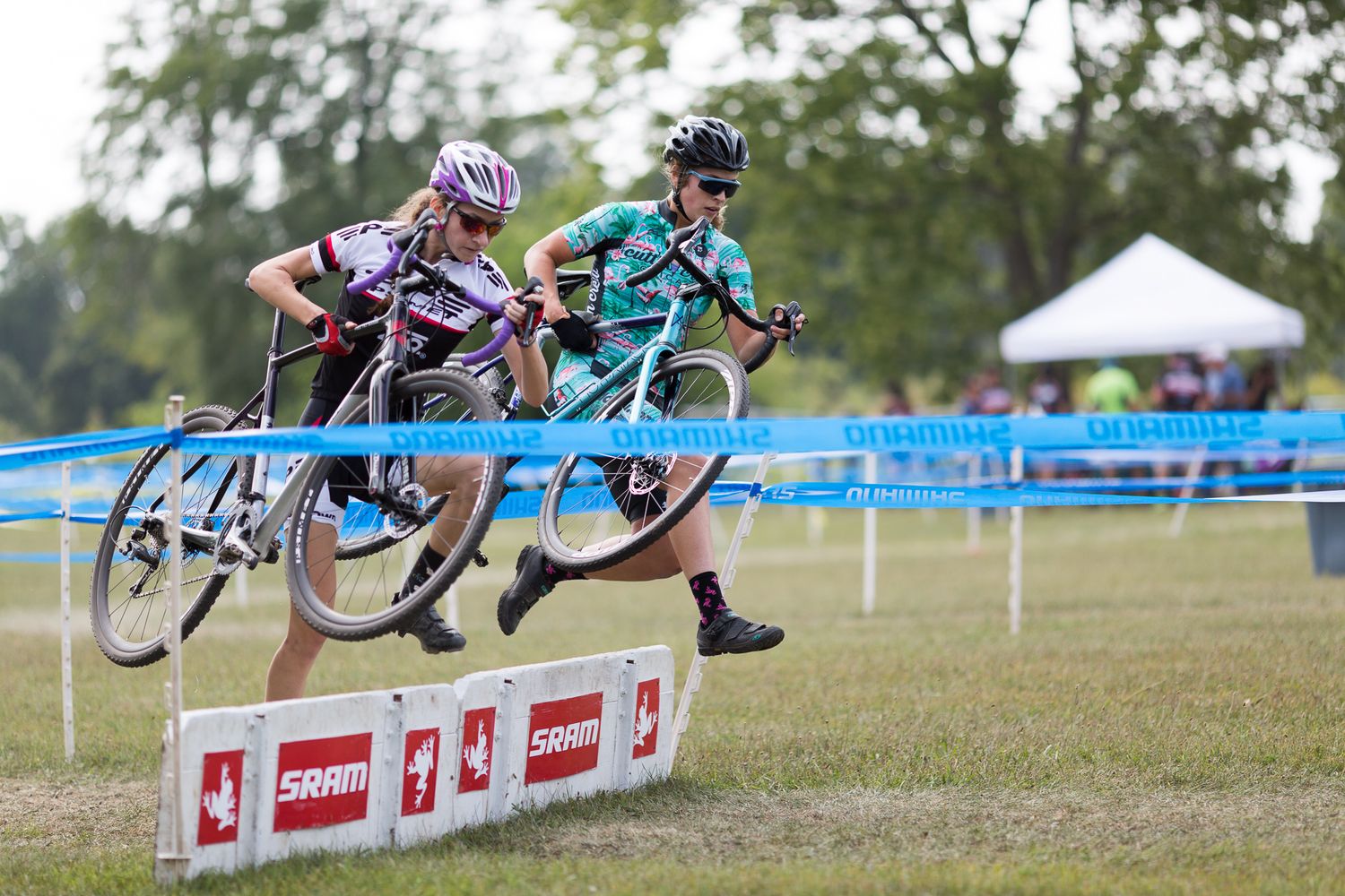Two young female cyclocross racers carry their bikes over SRAM hurdle barriers during a competitive race.