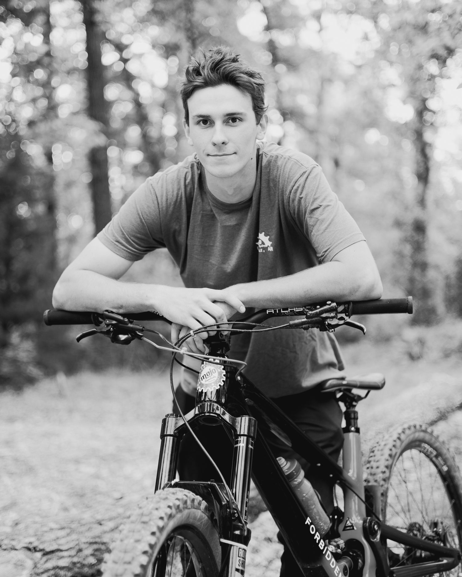 Black and white portrait of a young man posing with a mountain bike on a forest trail.