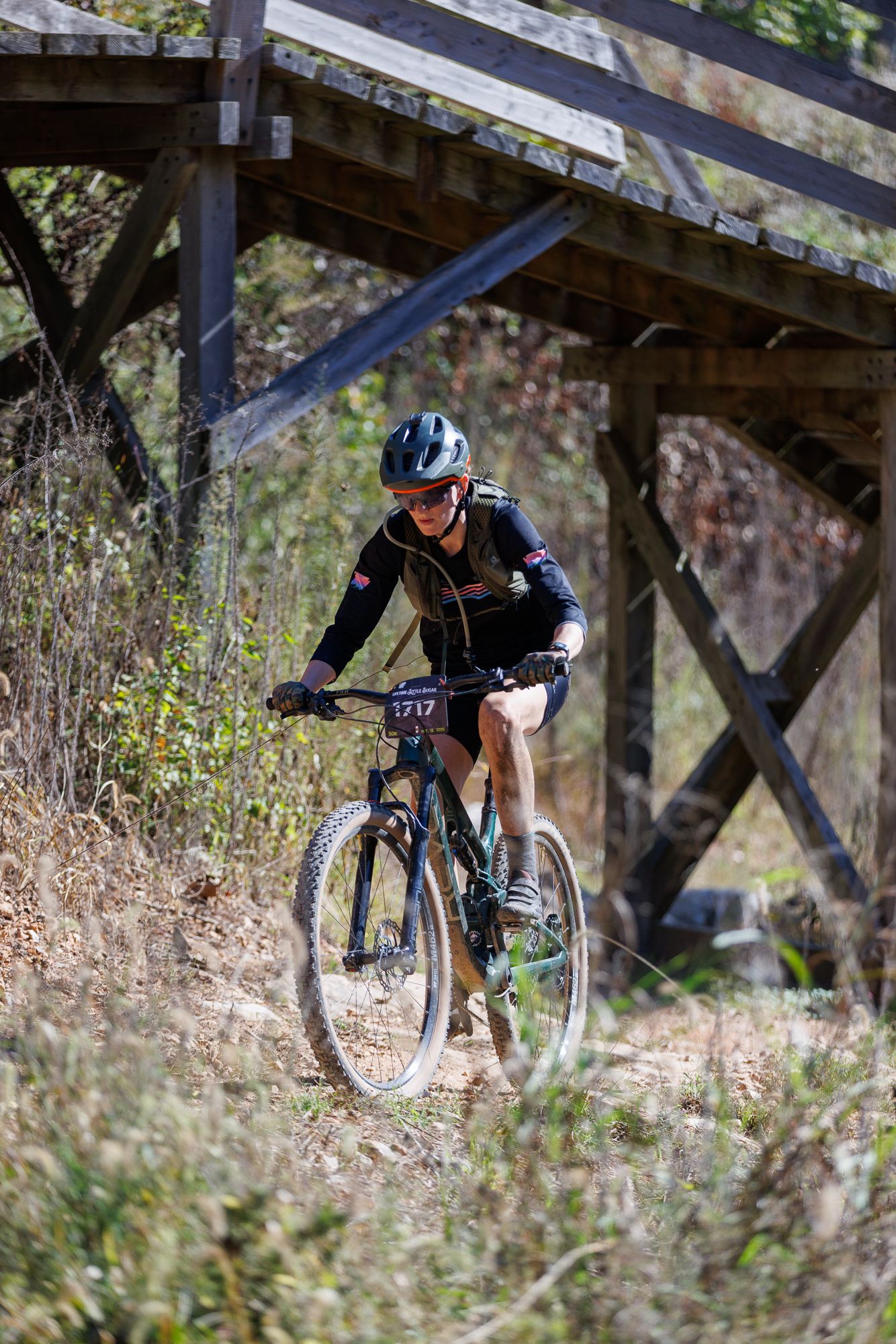 Mountain biker with race number 1717 rides dirt trail beneath rustic wooden bridge structure.