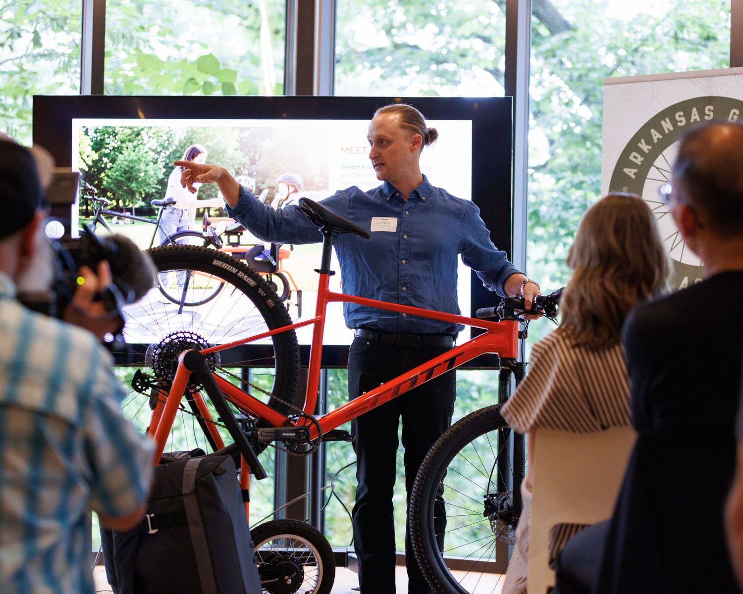 Man presenting a red bicycle to an audience at an Arkansas cycling event indoors.