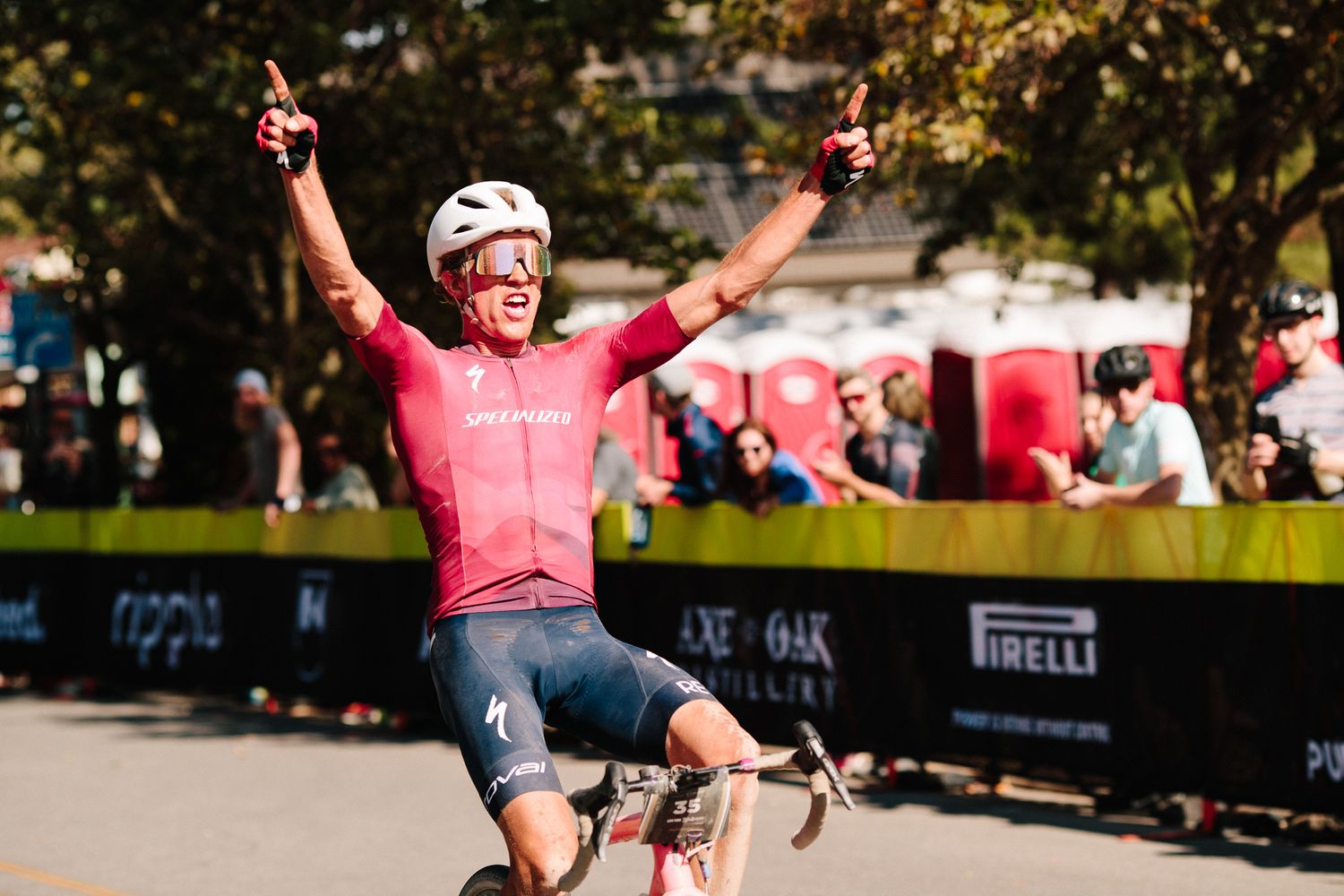 Cyclist in pink Specialized jersey celebrates a race victory with arms raised at finish line.