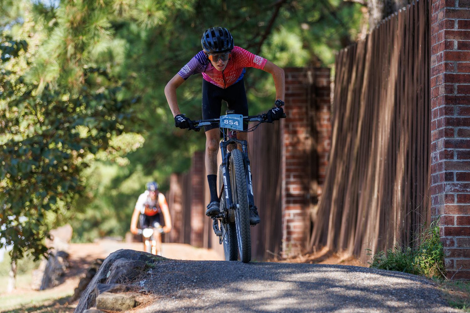 Mountain biker racing down a paved path near a brick wall during a competitive cycling event.