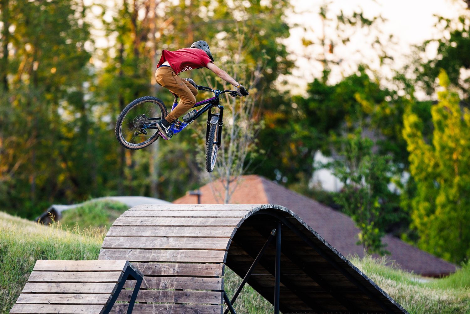 Mountain biker launching off a wooden ramp jump at an outdoor bike park at sunset.