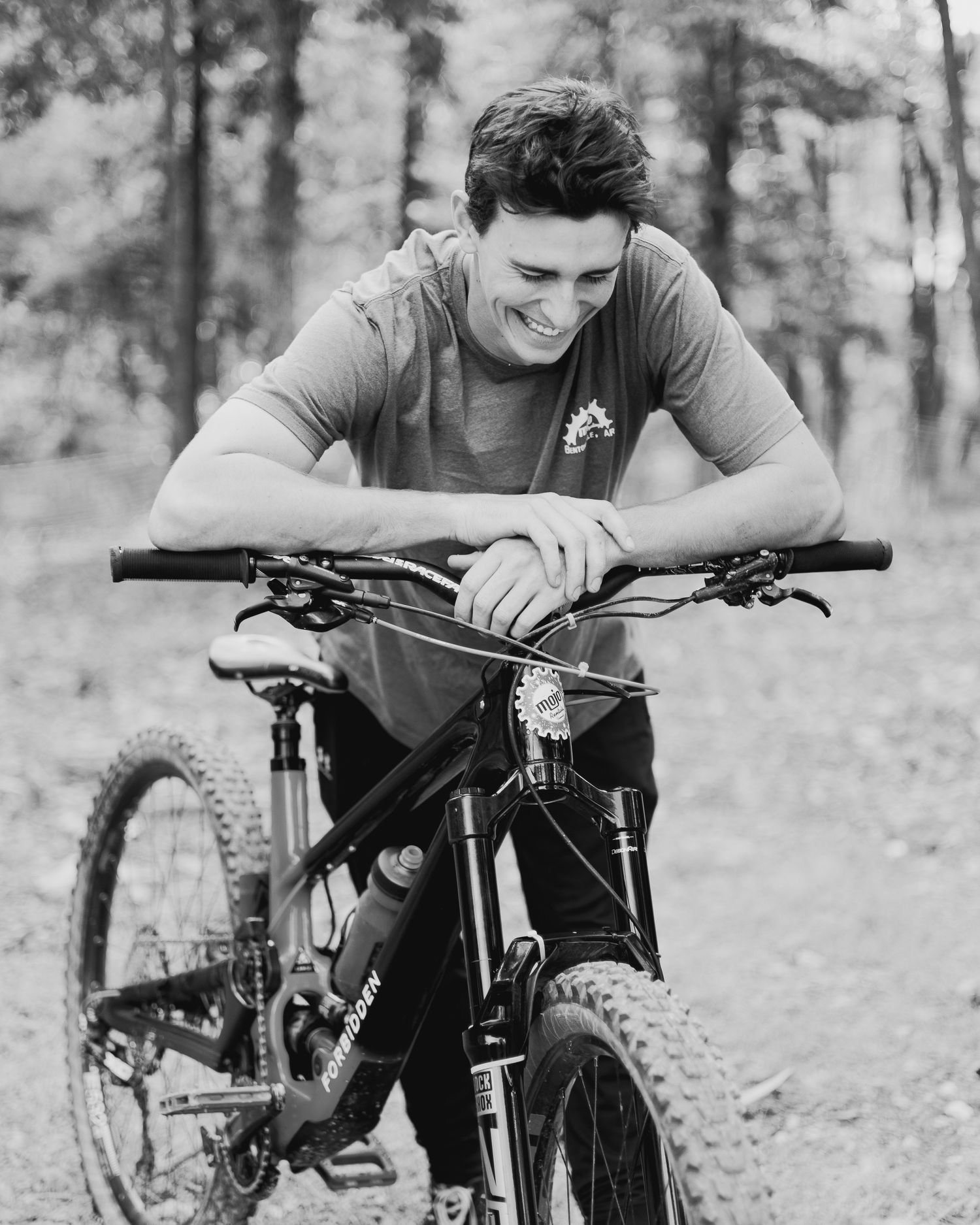 Black and white photo of a smiling young man leaning over a mountain bike in a forest setting.