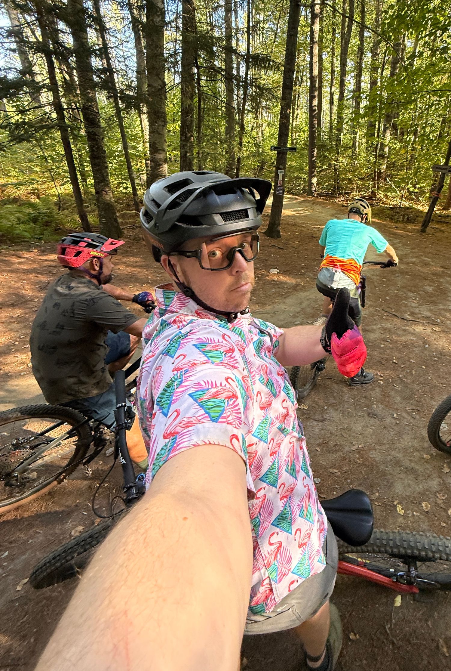 Mountain biker in flamingo print shirt and helmet takes selfie with group on sunny forest trail.
