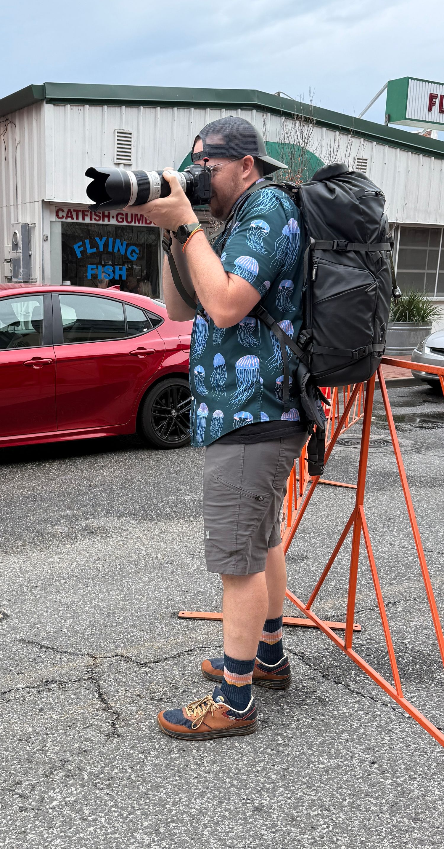 Photographer with large telephoto lens and backpack shooting outside Flying Fish market in urban setting.