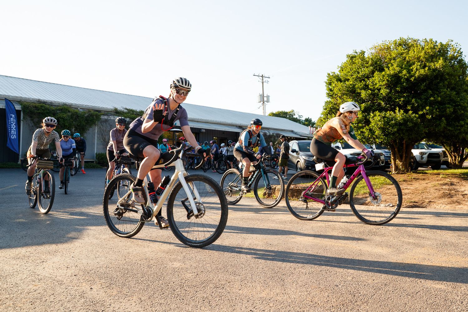 Group of cyclists in helmets start a gravel race on a paved road in warm sunny conditions.