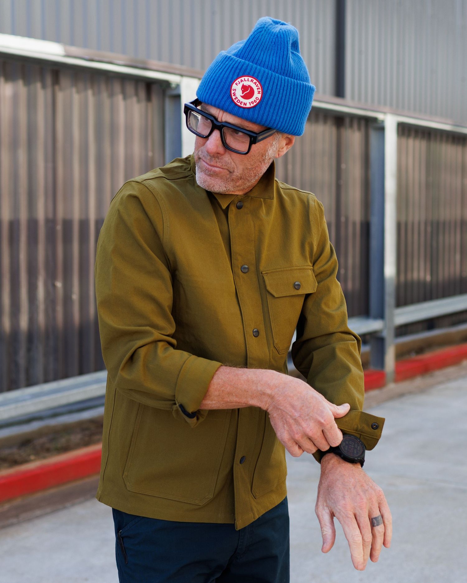 Man in olive jacket and blue beanie checking his watch outdoors.